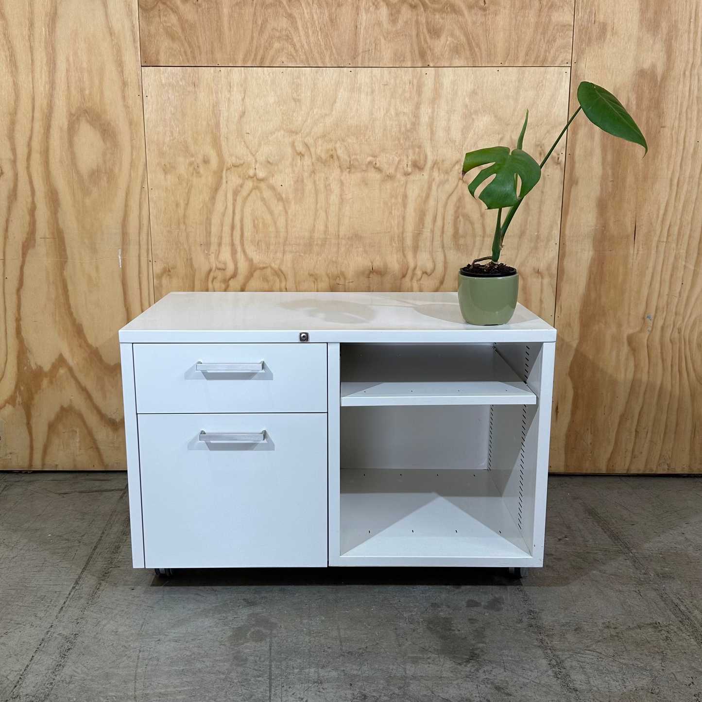 White office cabinet with drawers and a plant against a wooden wall.